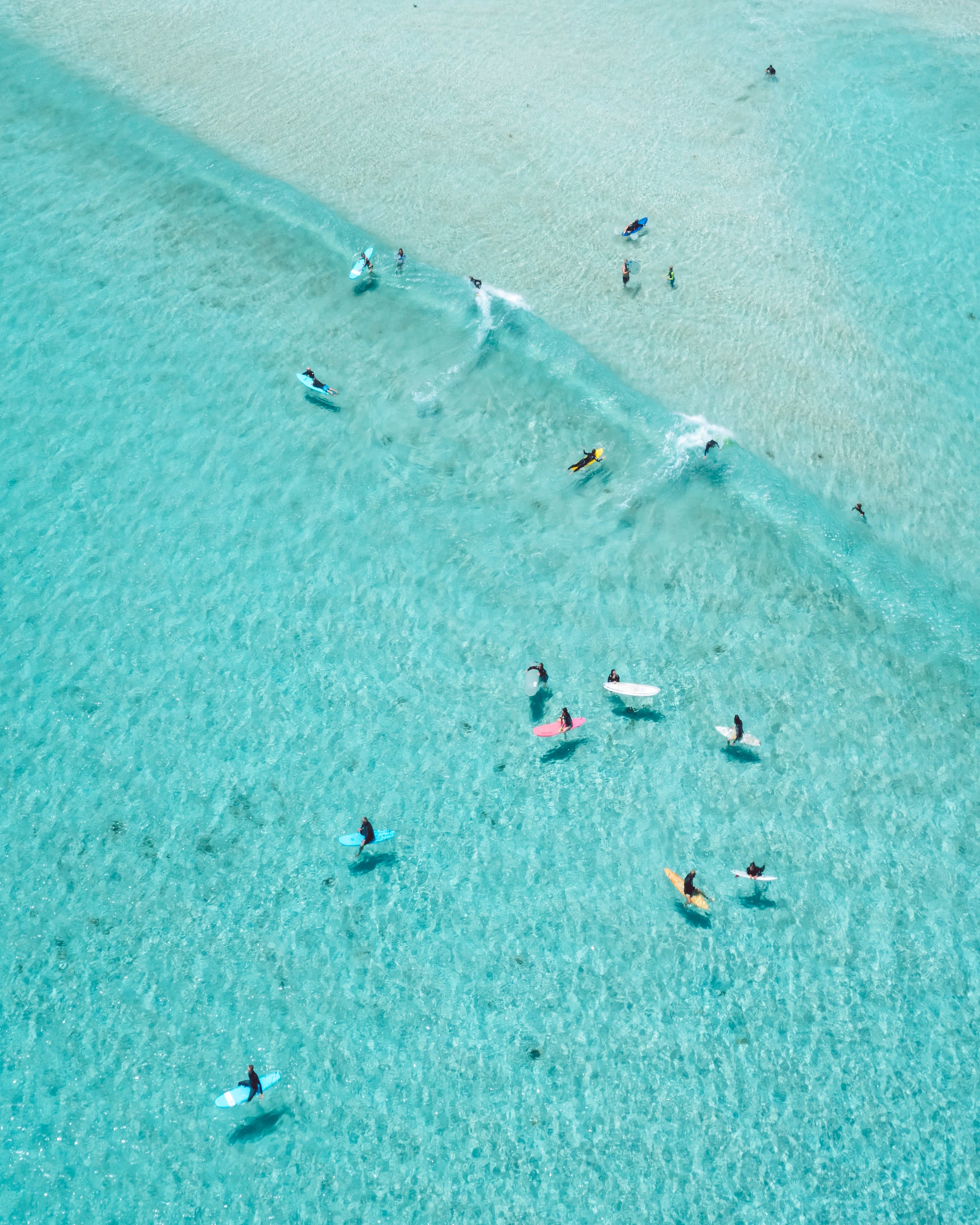 Aerial view of surfers at Smiths Beach, Western Australia, floating on vibrant turquoise water with visible sandy seabed. Brightly colored surfboards add pops of color, while gentle waves ripple across the scene.